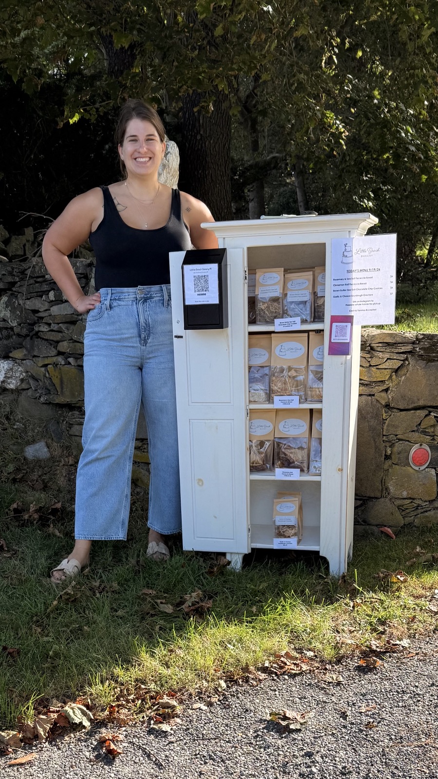 A smiling baker standing beside the Little Beach Bakery roadside stand, a white cabinet of bagged focaccia, cookies and crackers in front of a stone wall